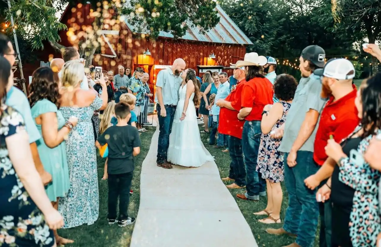 A newly married couple kisses at the end of the aisle as guests line the path, the barn behind them.