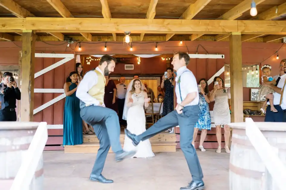 Two groomsmen dance under string lights on a covered porch while the bride and guests watch.