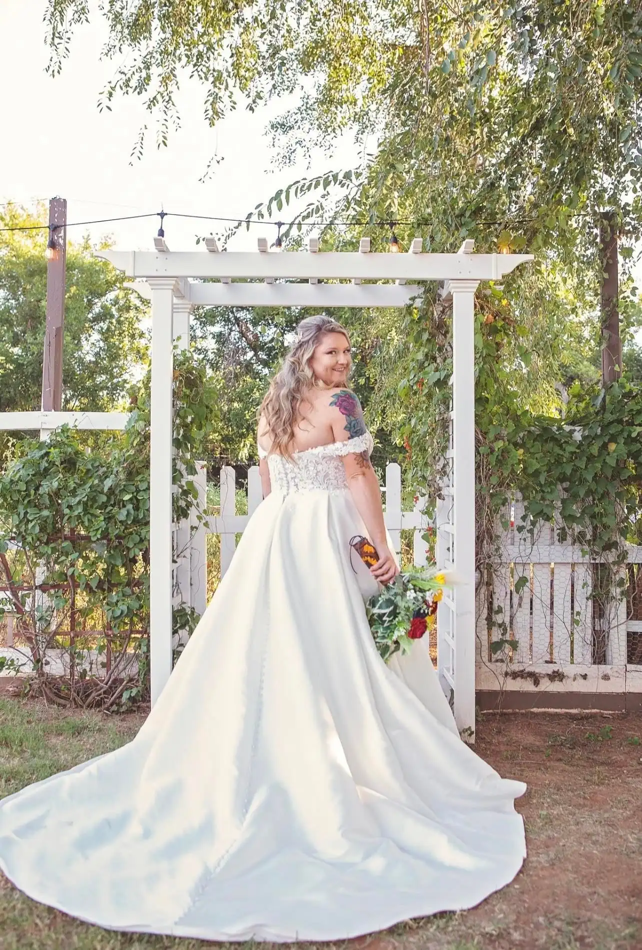 A bride in a satin gown looks back over her shoulder under a white garden arbor.