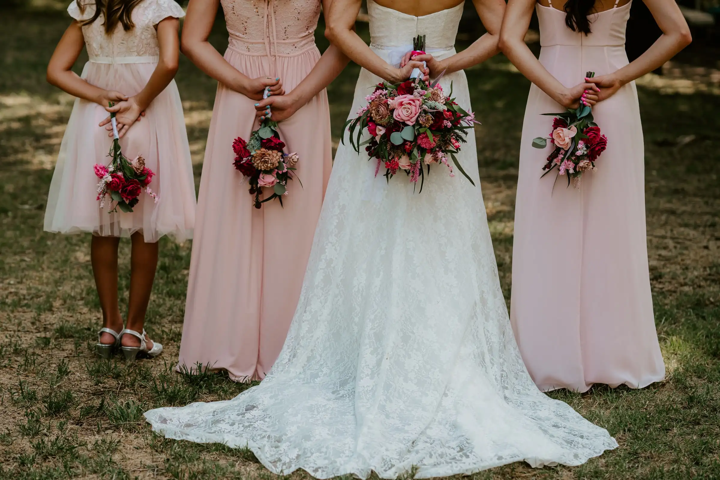 A bride and her bridesmaids in blush dresses hold bouquets behind their backs.