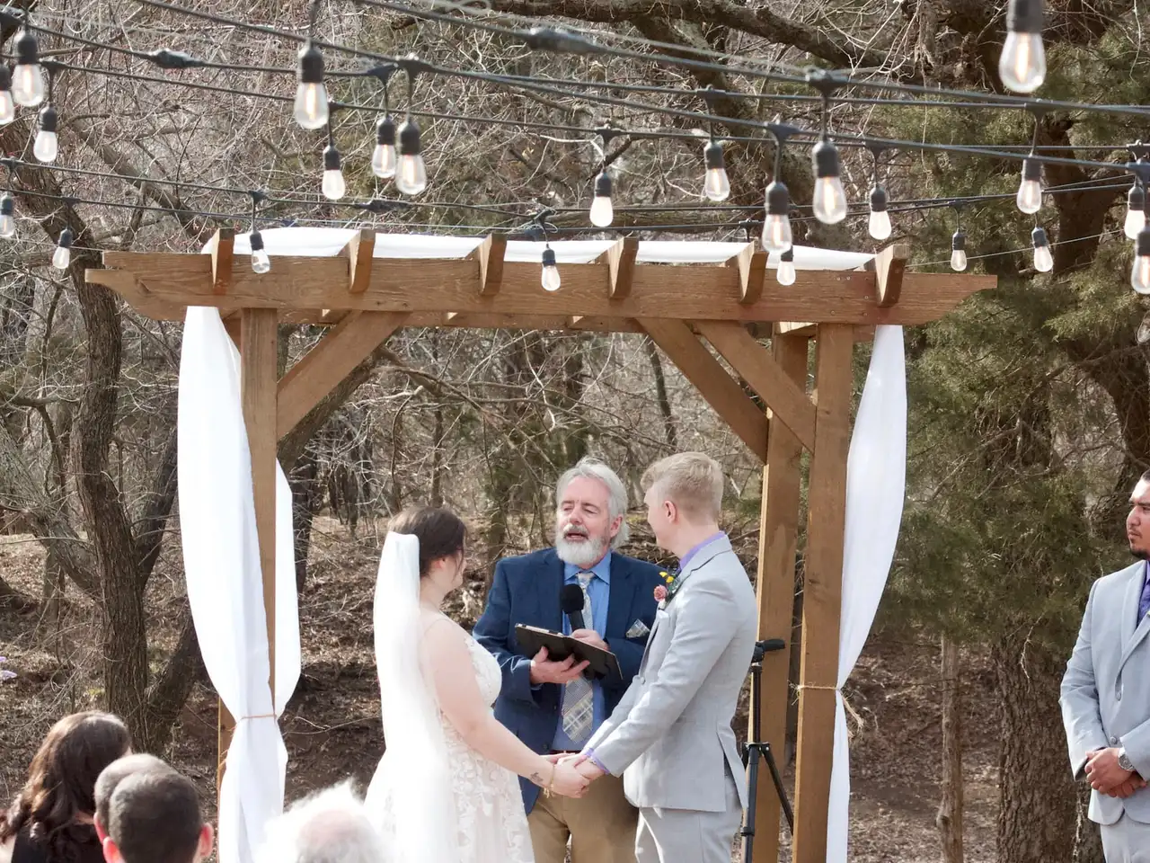 A ceremony under a wooden arbor with white drapes and globe string lights, guests seated on white chairs.