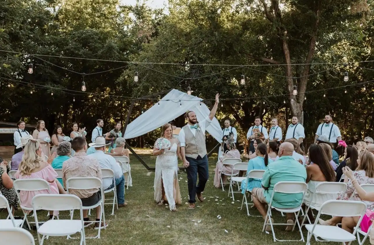 Guests celebrate as a couple walks back up the aisle under string lights and trees.