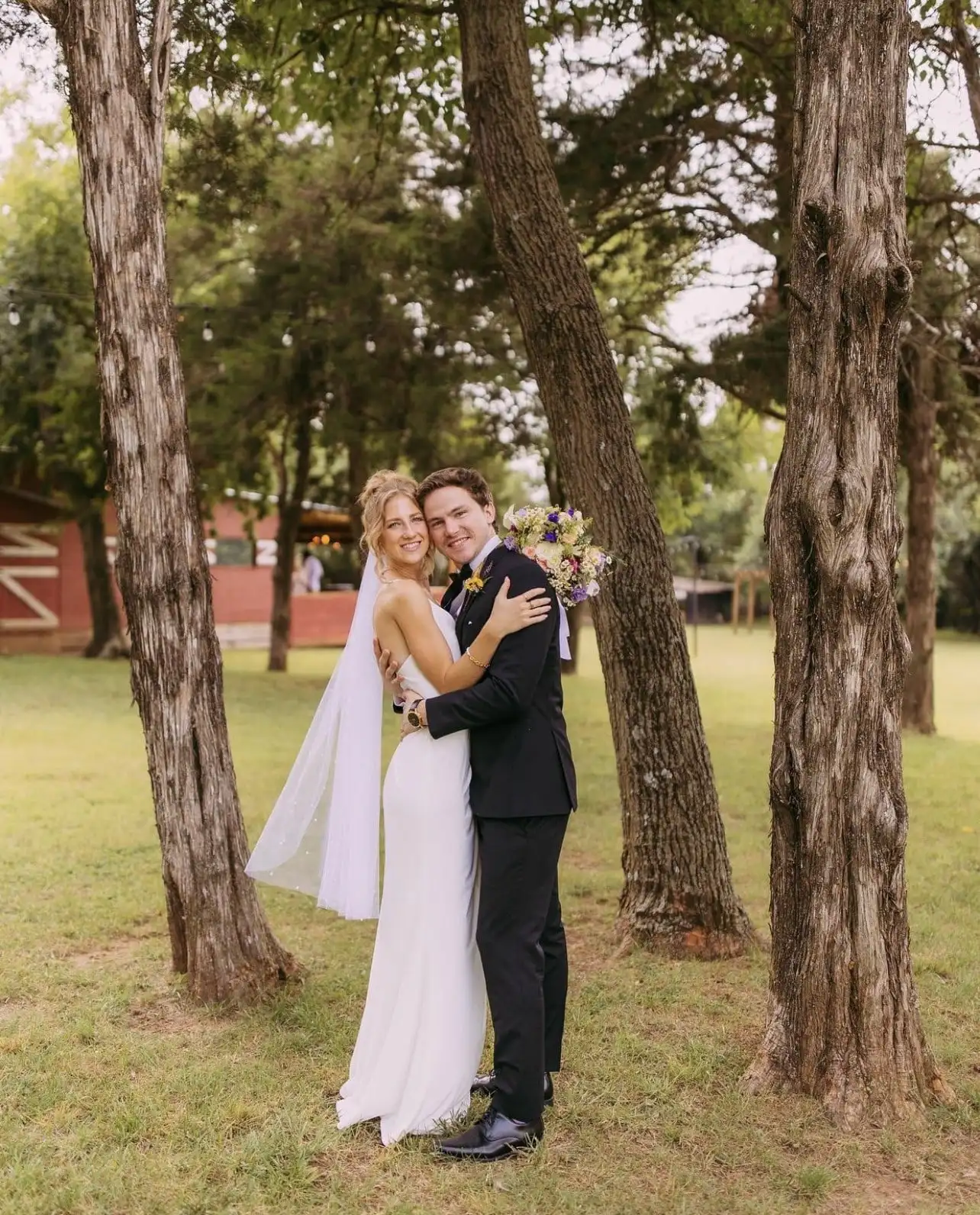 A bride and groom embrace between tall tree trunks on the lawn, the red barn behind them.