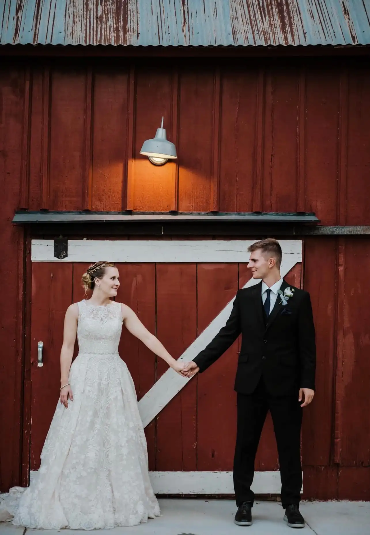 A bride and groom hold hands in front of the red barn's white-trimmed sliding door.
