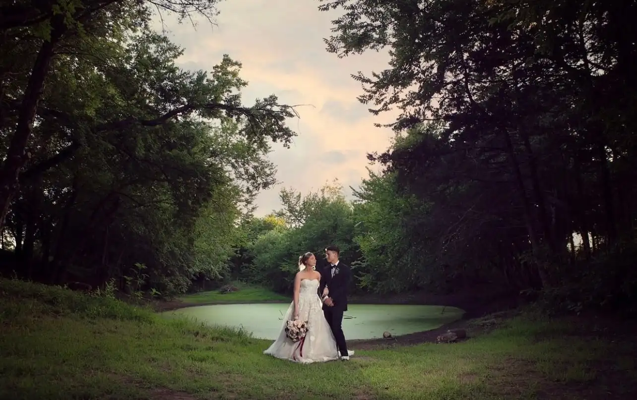 A bride and groom stand by the pond at dusk, framed by dark trees.