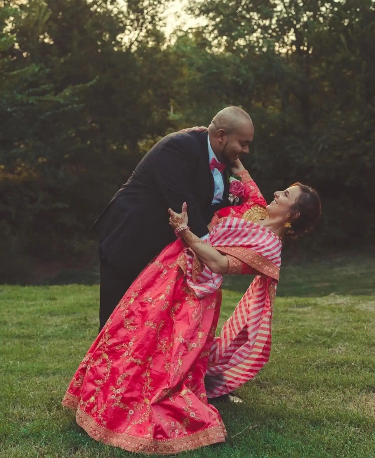A groom dips his bride, who wears a red and pink embroidered dress, on the lawn.