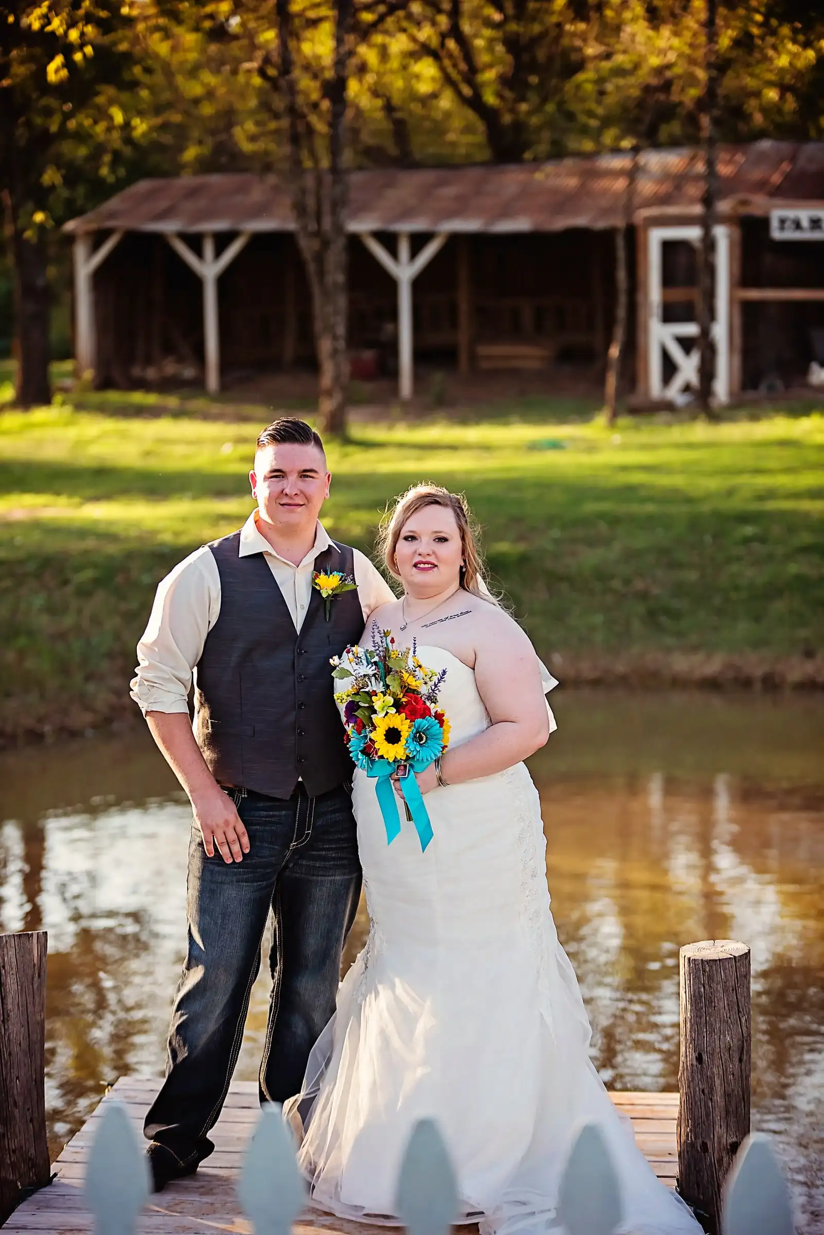 A bride with a sunflower bouquet and her groom stand on the dock by the pond.