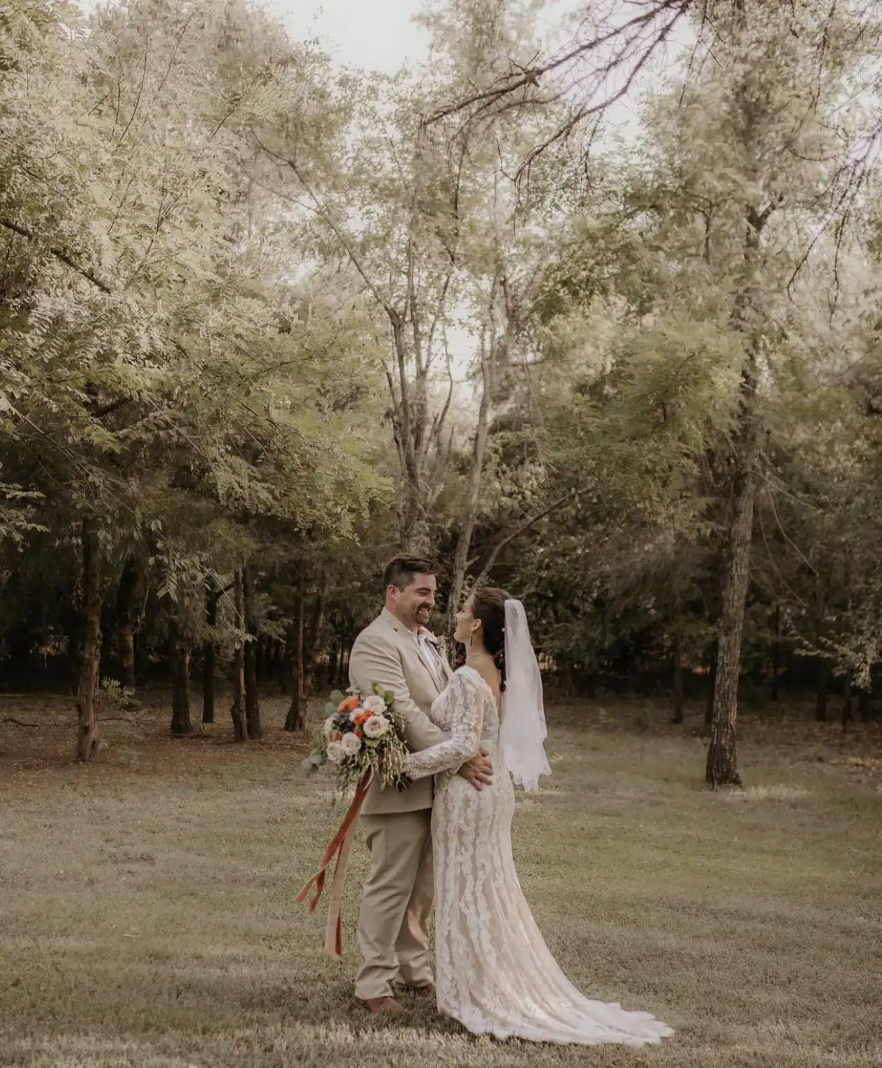 A bride and groom smile at each other in a grove of trees.