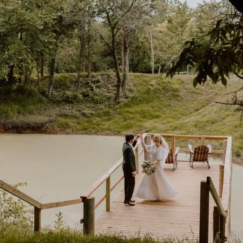 A groom spins his bride on a wooden dock over the pond.
