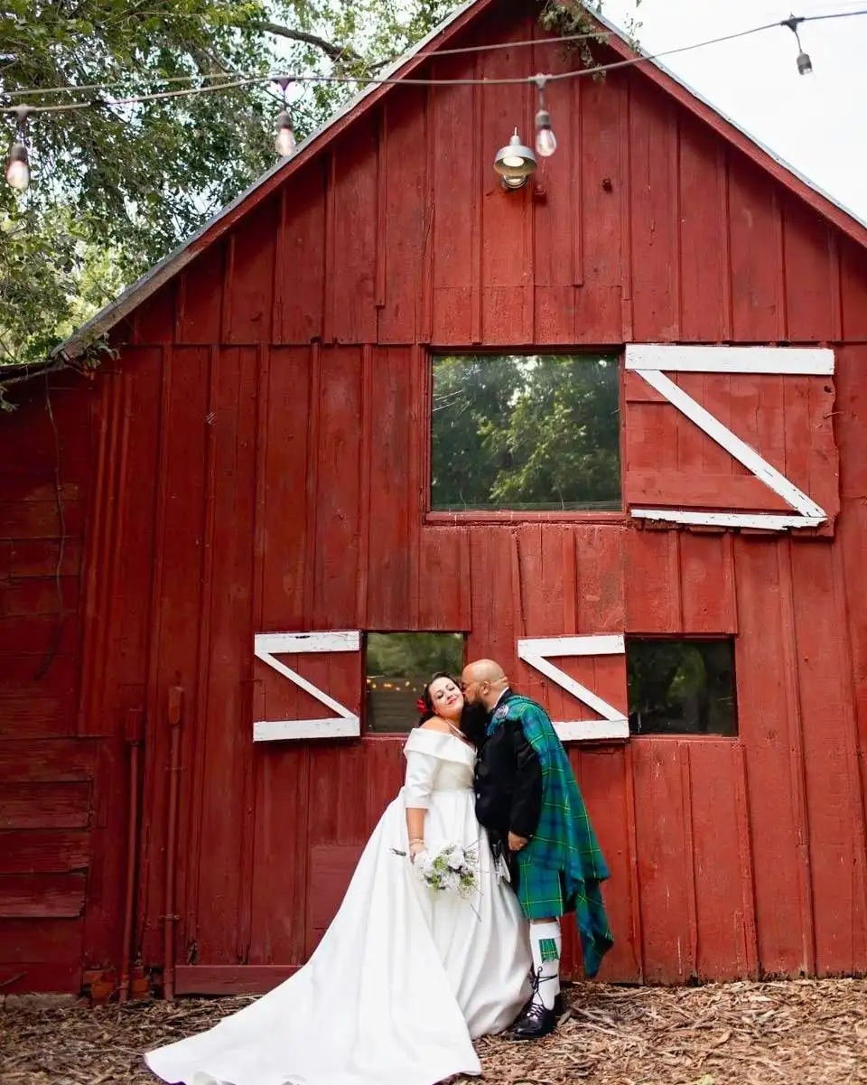 A groom in a green tartan kilt kisses his bride against the side of a red barn.