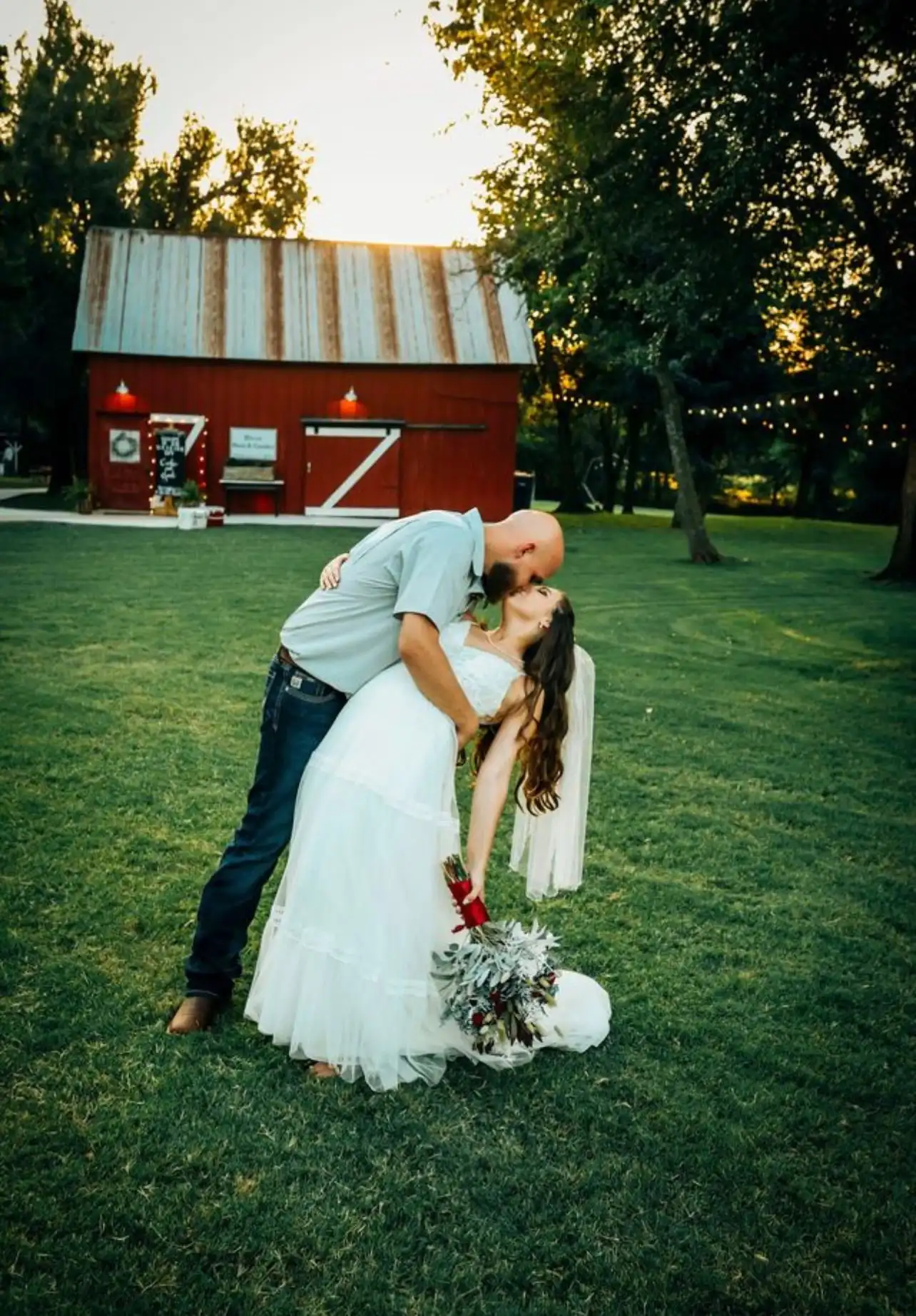 A groom dips his bride for a kiss on the lawn in front of the red barn at sunset.
