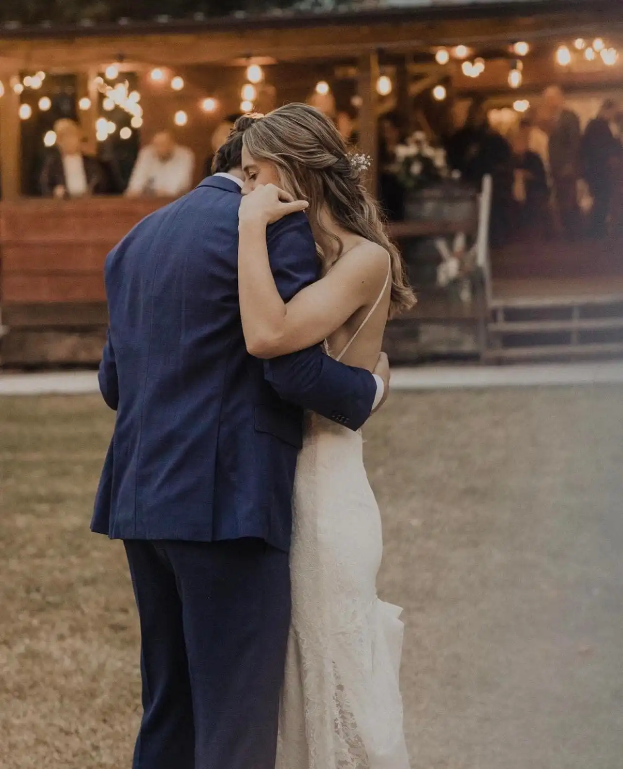 A couple holds each other close during their first dance, string lights glowing behind them.