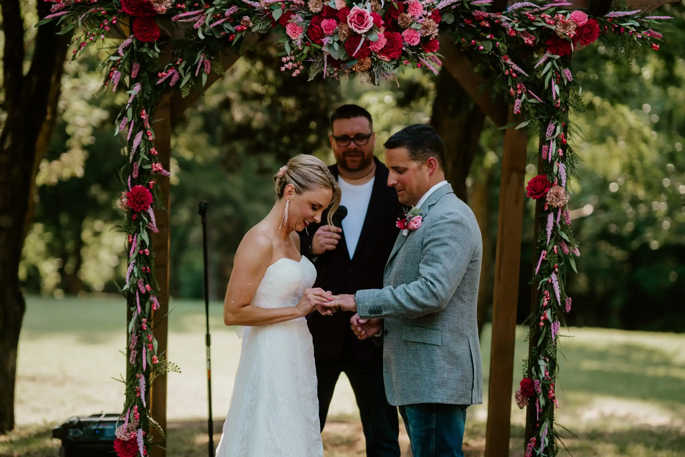 A couple exchanges rings under an arbor wrapped in red and pink flowers.