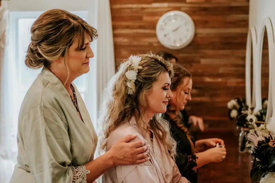 A bride in a blush robe gets ready while a woman rests a hand on her shoulder.