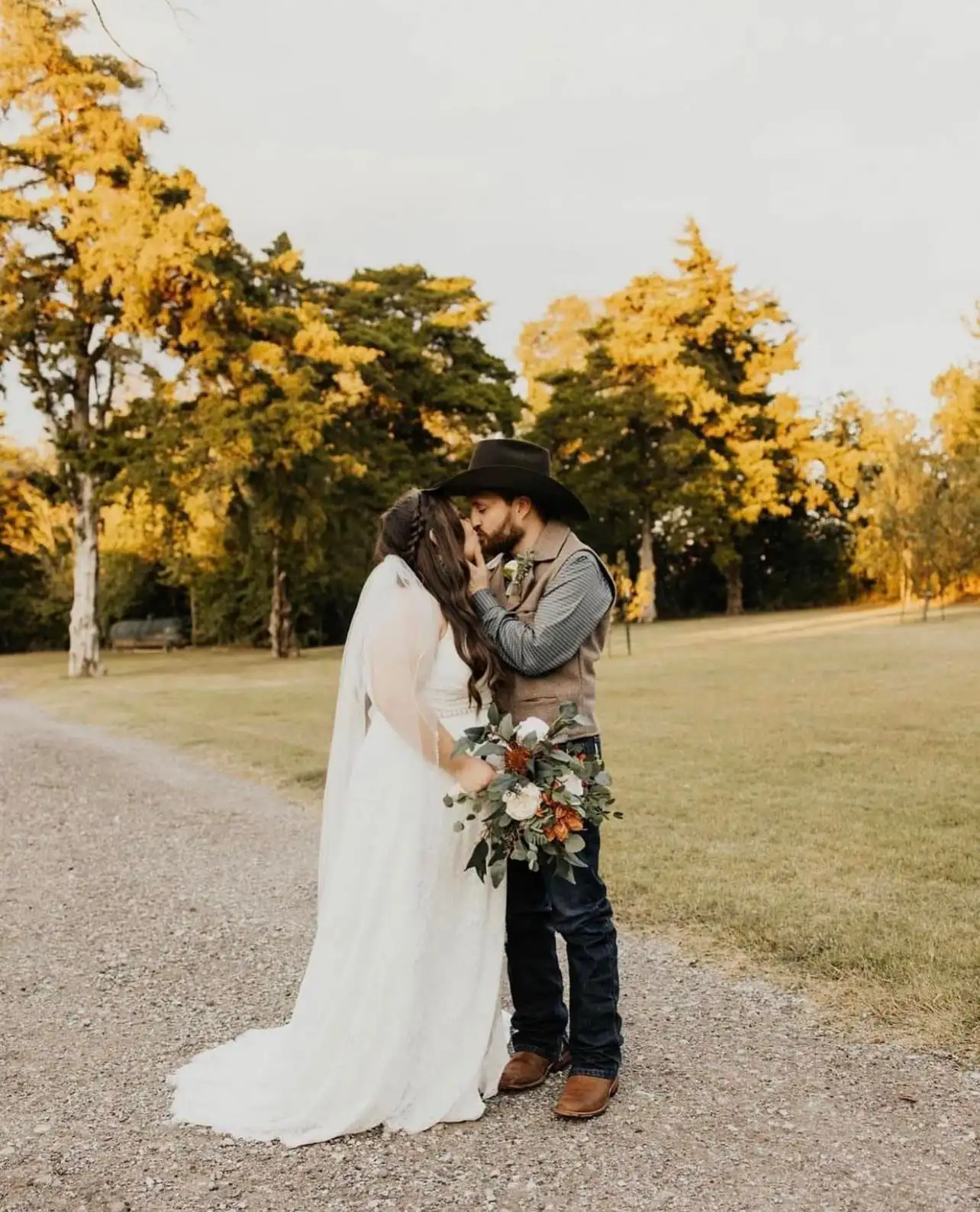 A groom in a cowboy hat kisses his bride on the gravel drive among fall trees.