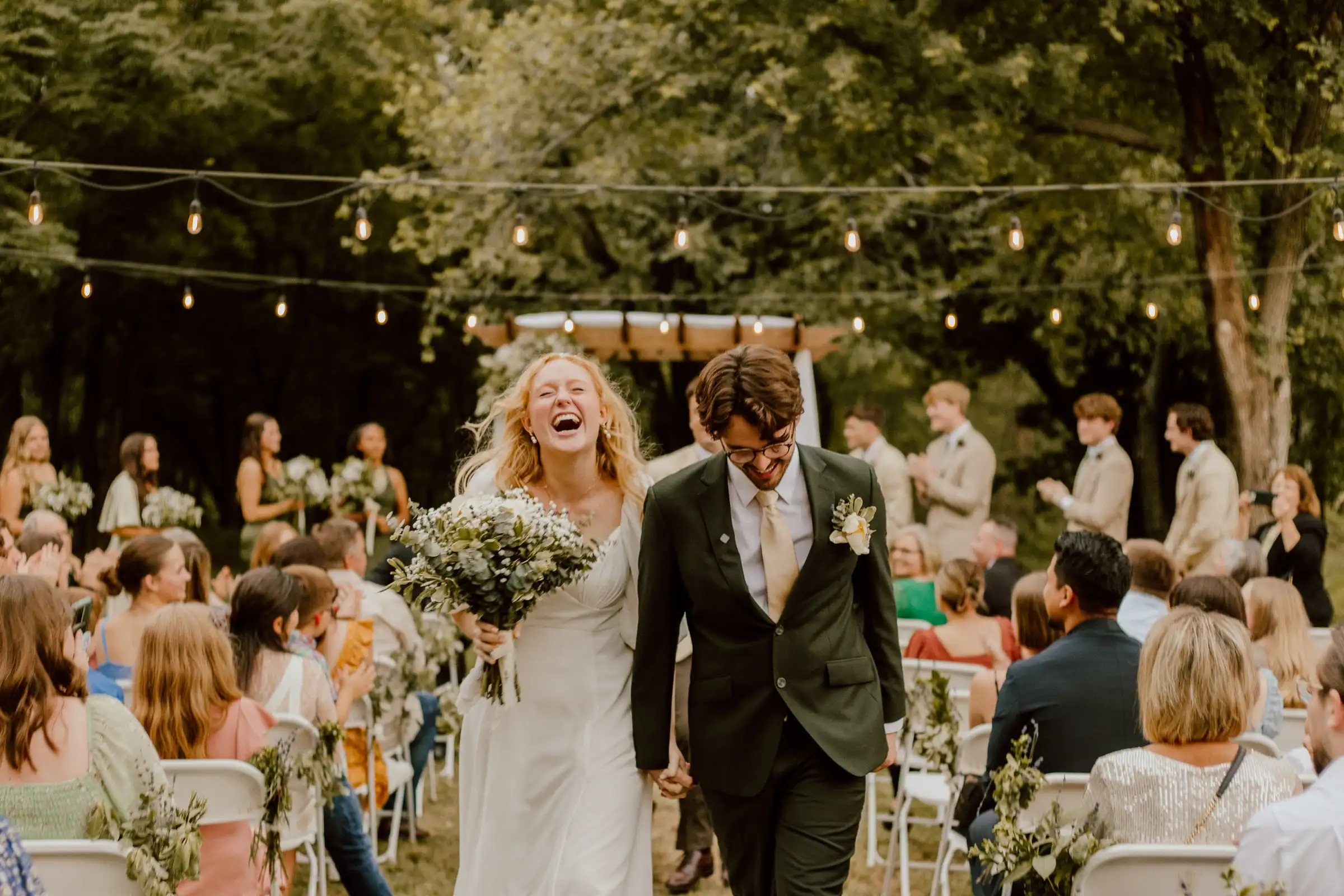 A laughing bride and groom walk back up the aisle under string lights as guests stand and cheer.
