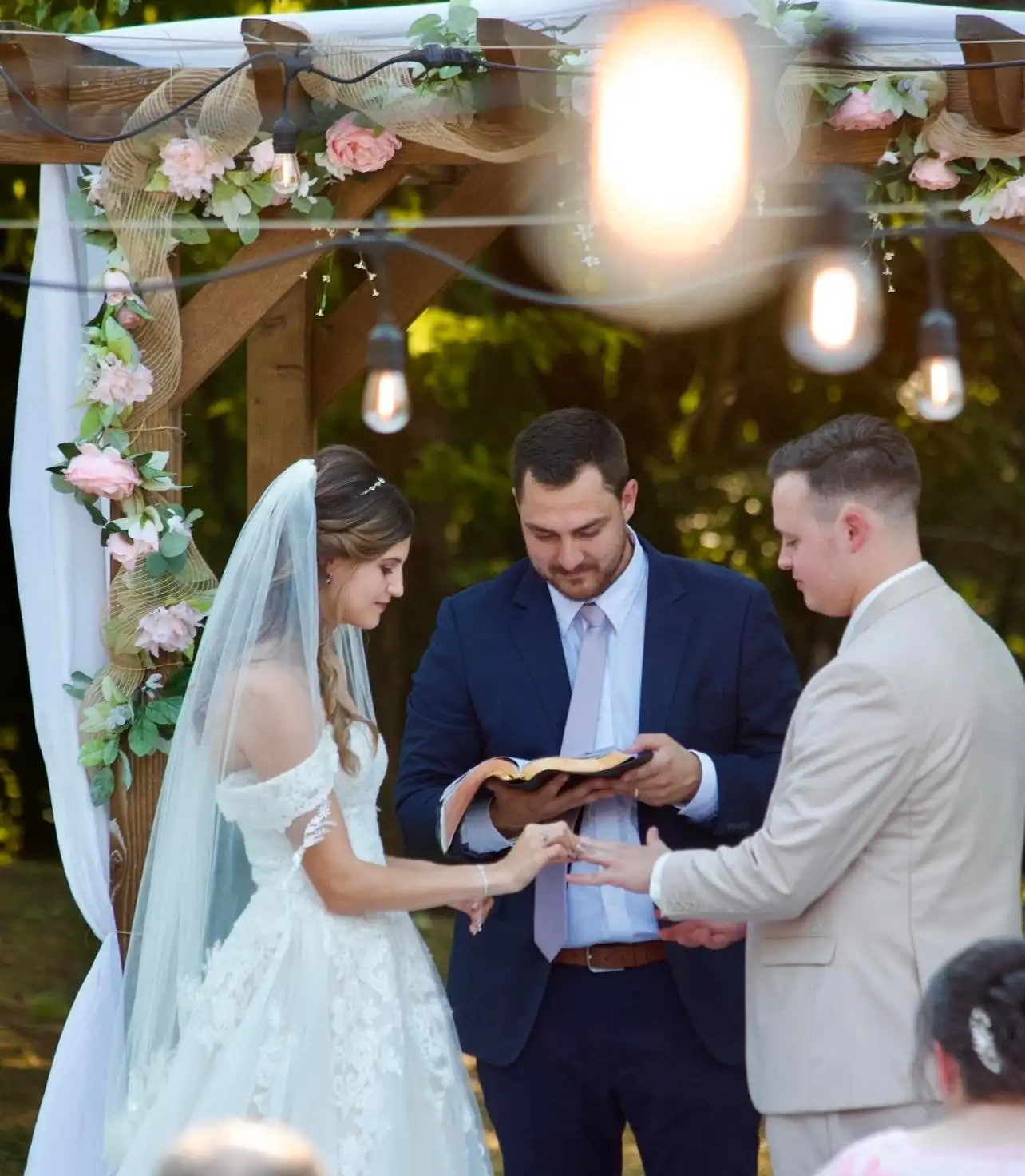 A couple exchanges rings under a wooden arbor hung with pink flowers and globe lights.