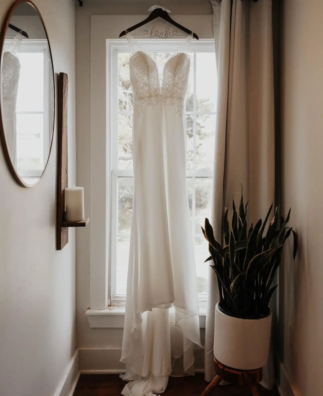 A wedding dress hangs in a bright window beside a round mirror.
