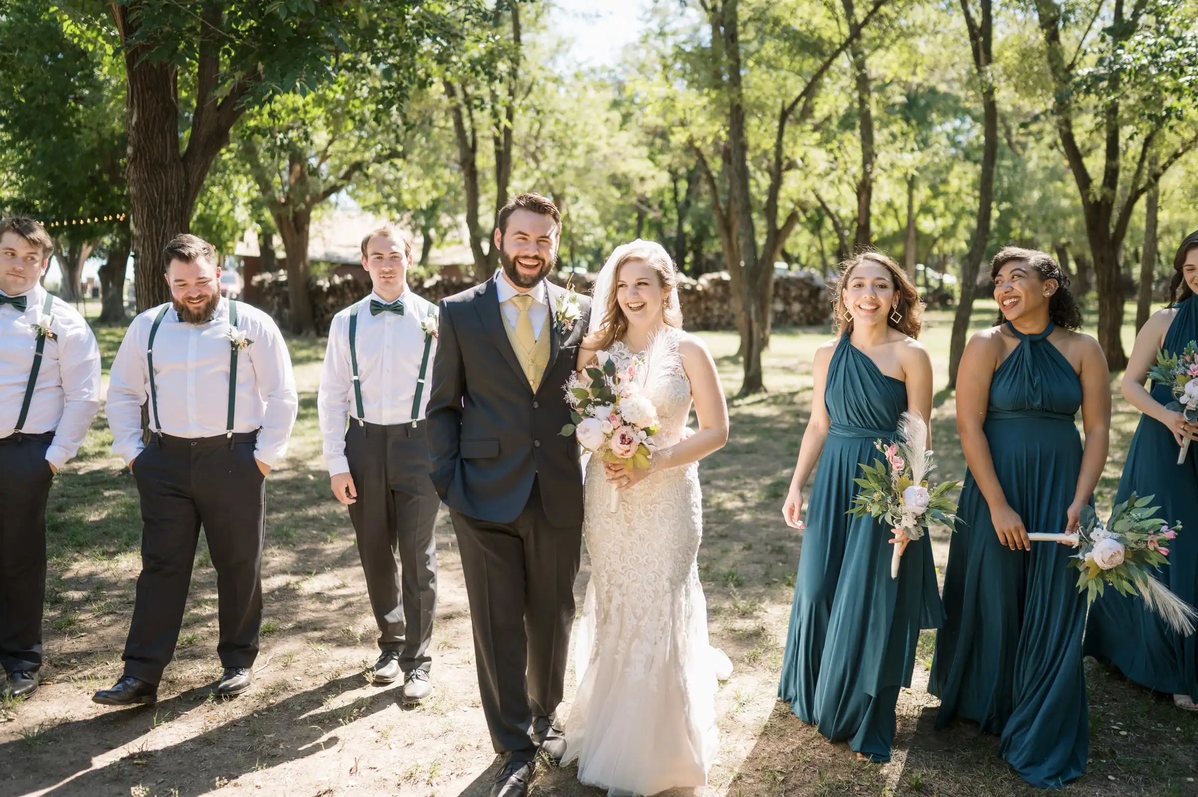 A bride and groom with their wedding party, bridesmaids in teal and groomsmen in suspenders, among the trees.